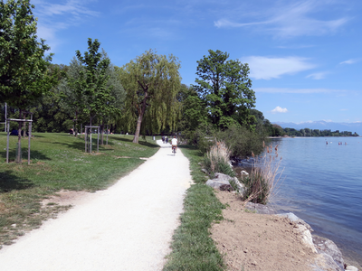 le chemin pédestre de la rive, avec le lac et ses enrochements actuels à droite, un jour de très beau temps. Un vélo s'éloigne. 