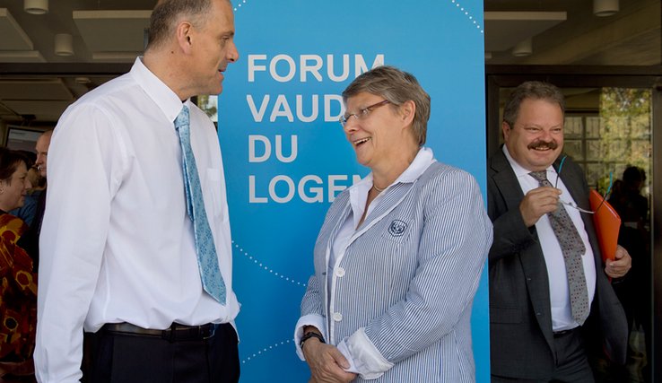 Les trois personnages posent devant un panneau "Forum vaudois du logement".