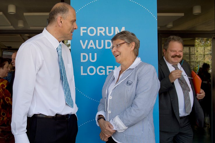 Les trois personnages posent devant un panneau "Forum vaudois du logement".