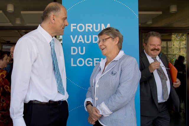 Les trois personnages posent devant un panneau "Forum vaudois du logement".