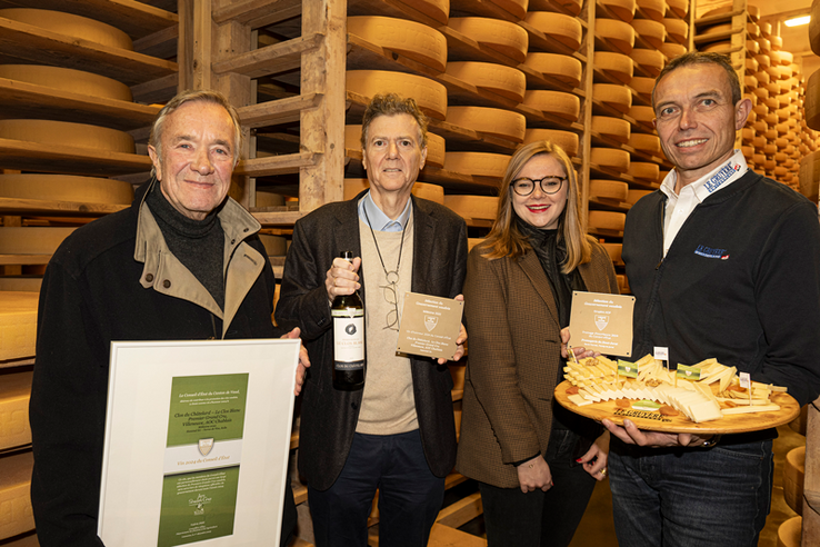 Les personnes posent avec diplômes, bouteille et plateau de fromage dans la cave de la fromagerie à Peney-le-Jorat. Derrière, les rayonnages pleins de meules de gruyère.