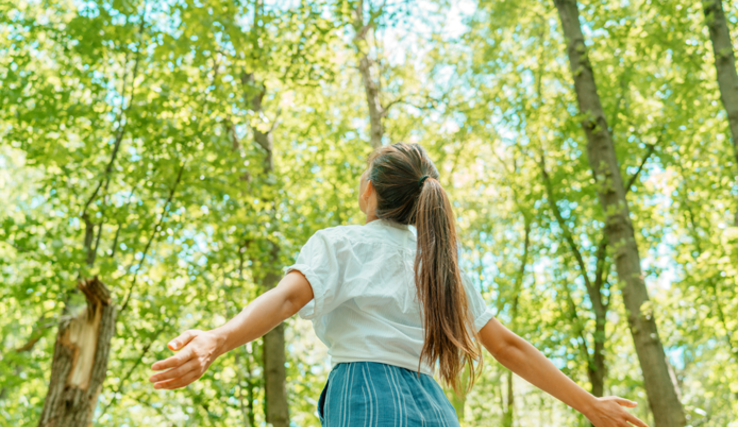 une jeune femme, de dos, bras écarté, dans une forêt. Au-dessus des arbres, le ciel bleu