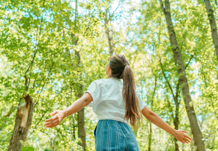 une jeune femme, de dos, bras écarté, dans une forêt. Au-dessus des arbres, le ciel bleu