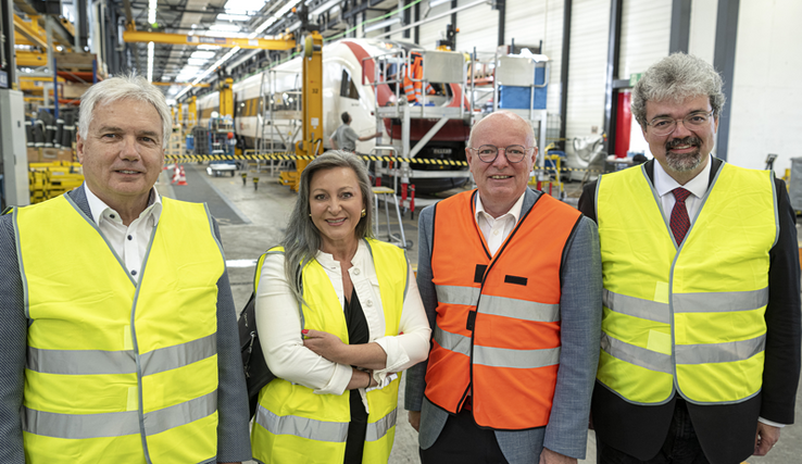 Les quatre personnages posent dans une halle des ateliers CFF d'Yverdon, vêtus de gilets haute-visibilité.