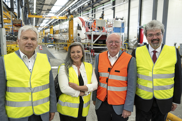 Les quatre personnages posent dans une halle des ateliers CFF d'Yverdon, vêtus de gilets haute-visibilité.