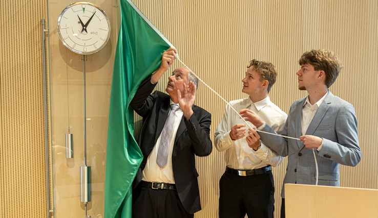 Les personnes sont au-dessous de l'horloge, les apprentis tirent sur sur une ficelle qui retire le voile qui masquait l'horloge. Le cabinet de l'horloge est en verre (ou en matériel transparent). On voit les poids et le balancier au-dessous du cadran.