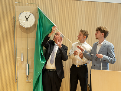 Les personnes sont au-dessous de l'horloge, les apprentis tirent sur sur une ficelle qui retire le voile qui masquait l'horloge. Le cabinet de l'horloge est en verre (ou en matériel transparent). On voit les poids et le balancier au-dessous du cadran.