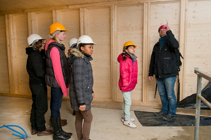 Un groupe de jeunes filles portant des casques de chantier face à un technicien, index dressé, qui semble leur donne des explications.