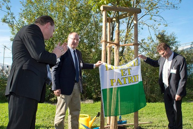 Monsieur Leuba est à gauche de l'image, il applaudit. Messieurs Maillard et Villiger se tiennent de part et d'autre de l'arbre. Ils tiennent un drapeau vaudois. Au sol, deux bêches et un arrosoir.