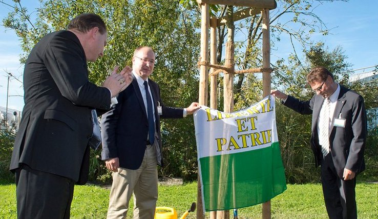 Monsieur Leuba est à gauche de l'image, il applaudit. Messieurs Maillard et Villiger se tiennent de part et d'autre de l'arbre. Ils tiennent un drapeau vaudois. Au sol, deux bêches et un arrosoir.