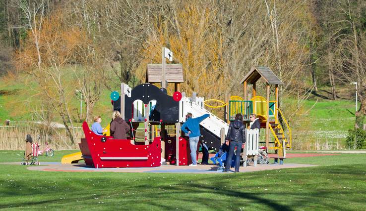 Enfants jouant dans une installation dans un parc public, sous le regard de quelques parents.