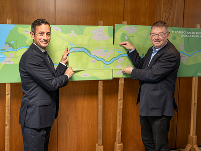 les deux personnages, debout devant une carte du projet, se serrent la main.