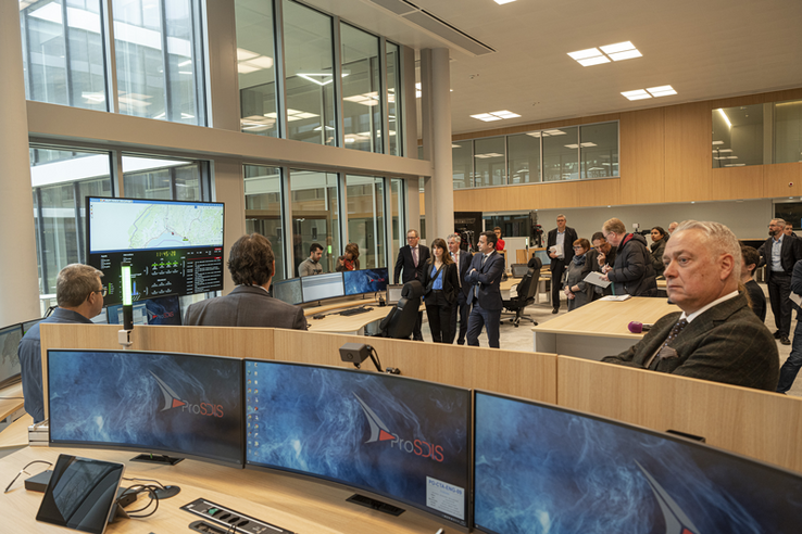 Photo lors de l'inauguration. Au 1er plan, un pupitre avec trois grands écrans. Les personnalités (la conseillère d'Etat Rebecca Ruiz et son collègue Vassilis Venizelos) sont derrière dans un grand espace vitré. A gauche, un écran pour le monitoring des alertes en temps réel. A droite de la photo, monsieur Froidevaux, chef de l'Etat-major cantonal de conduite.