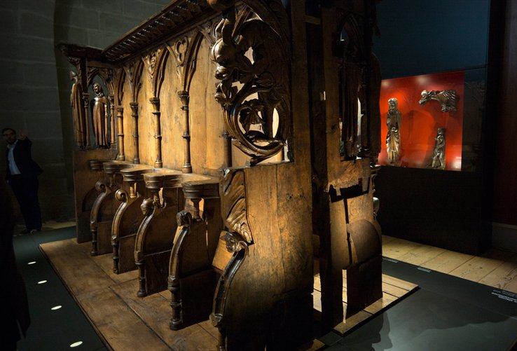 Les stalles de bois sculpté sous un éclairage calculé. Au fond, dans une vitrine, d'autres objets anciens en bois sculpté.