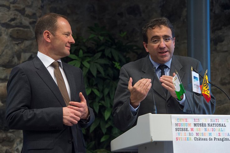 Messieurs Dal Busco et Broulis. Derrière un pupitre, Pascal Broulis parle en joignant les mains.