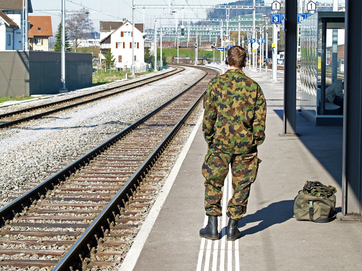 Un soldat de dos, en tenue de camouflage, attend le train, seul sur un quai de gare.
