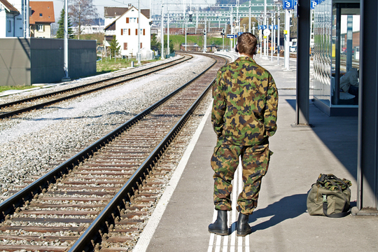 Un soldat de dos, en tenue de camouflage, attend le train, seul sur un quai de gare.