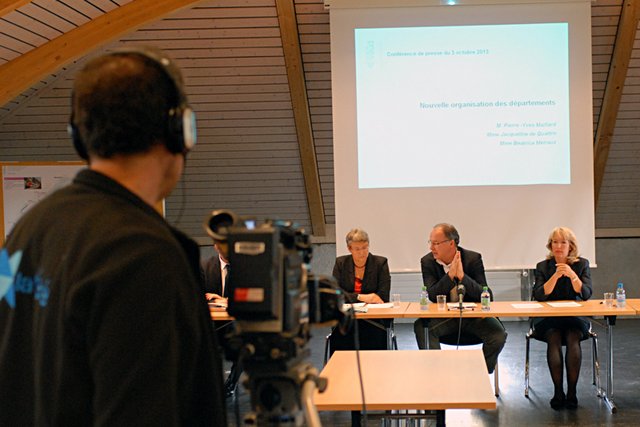 Les trois conseillers d'Etat derrière une table en conférence de presse. En arrière-plan, un écran qui sert aux projections de documents. Au premier plan, le bras d'un cameraman de la TV.