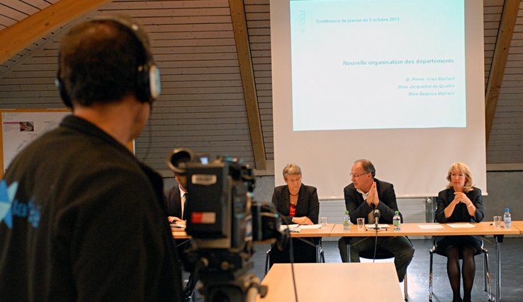 Les trois conseillers d'Etat derrière une table en conférence de presse. En arrière-plan, un écran qui sert aux projections de documents. Au premier plan, le bras d'un cameraman de la TV.