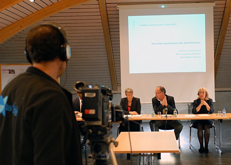Les trois conseillers d'Etat derrière une table en conférence de presse. En arrière-plan, un écran qui sert aux projections de documents. Au premier plan, le bras d'un cameraman de la TV.