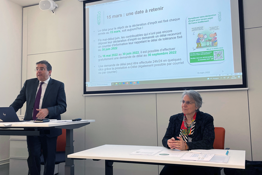 Les deux personnages en conférence de presse, devant un écran.