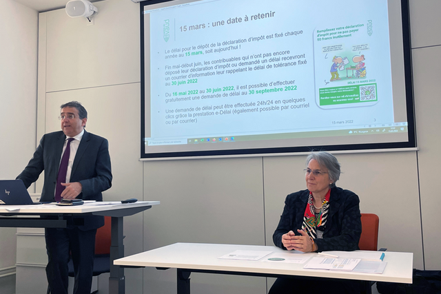 Les deux personnages en conférence de presse, devant un écran.