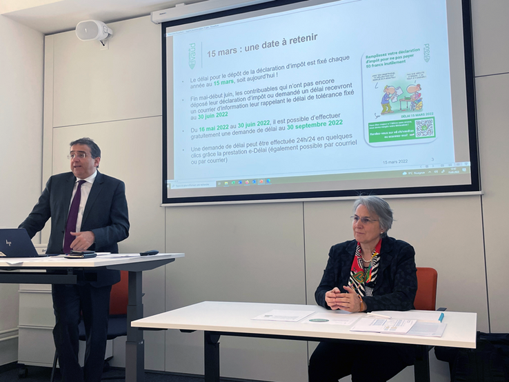 Les deux personnages en conférence de presse, devant un écran.