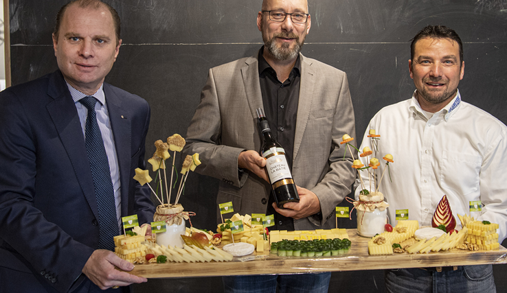 Les trois personnes posent, tenant une planche avec un assortiment de fromages. Monsieur Pasche tient une bouteille de vin