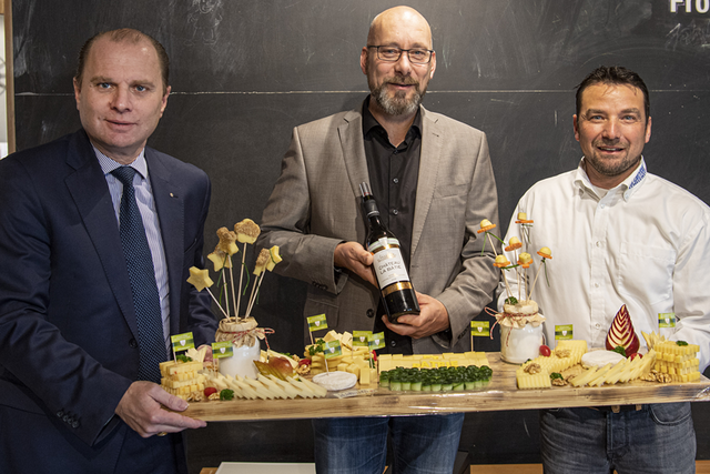 Les trois personnes posent, tenant une planche avec un assortiment de fromages. Monsieur Pasche tient une bouteille de vin