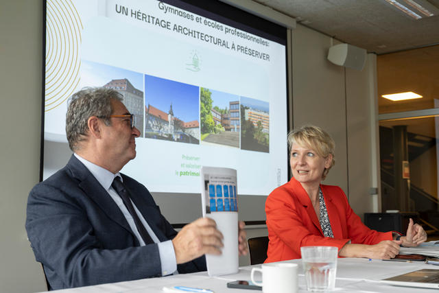 Les deux personnages durant la conférence de presse, assis à une table, devant un écran de projection. Ils sont en train d'échanger un regard.