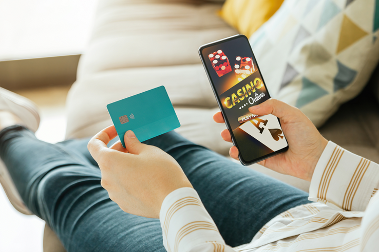 Une personnes allongée sur un canapé tient une carte de crédit dans la main gauche et dans la droite, un téléphone portable qui affiche un jeu de casino en ligne.