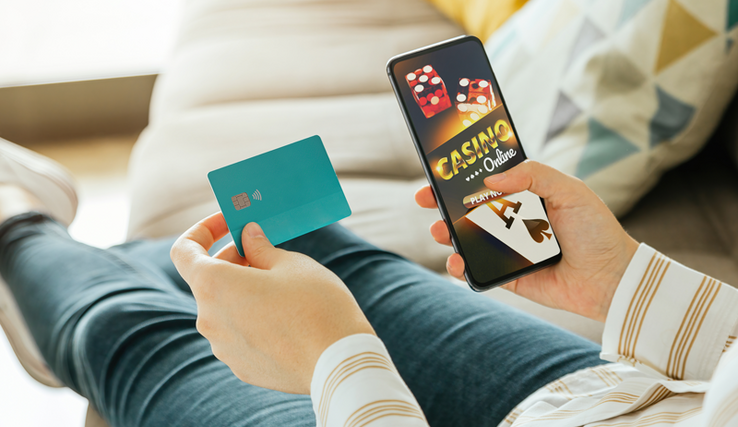 Une personnes allongée sur un canapé tient une carte de crédit dans la main gauche et dans la droite, un téléphone portable qui affiche un jeu de casino en ligne.