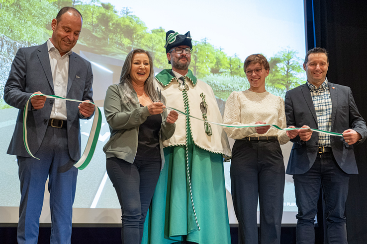 Les personnages posent sur une scène devant un écran qui montre le futur tunnel qui sera construit au-dessous de Sainte-Croix