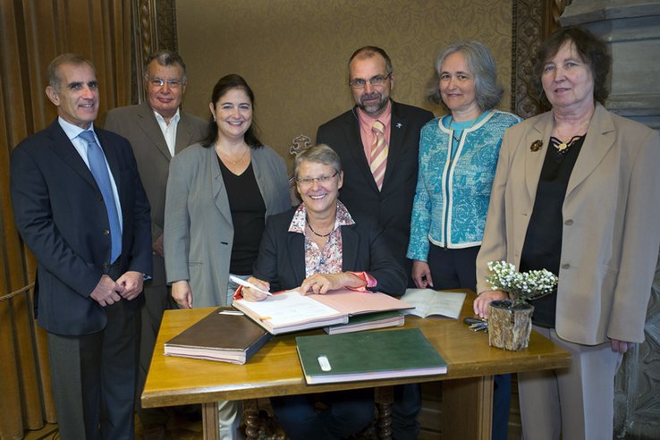 Les personnages posent derrière une table où est posé un registre. Madame Métraux est assise et tient un stylo au-dessus du registre.