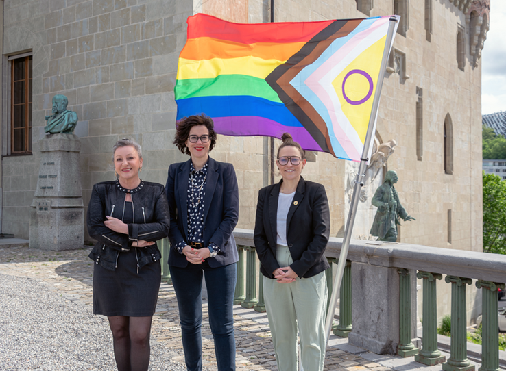 Les trois personnes posent devant le drapeau multicolore. Derrière, les façades du château Saint-Maire et la statue du major Davel.
