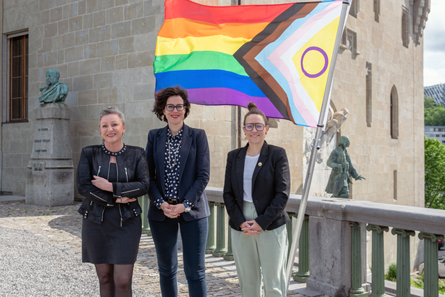 Les trois personnes posent devant le drapeau multicolore. Derrière, les façades du château Saint-Maire et la statue du major Davel.