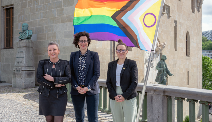 Les trois personnes posent devant le drapeau multicolore. Derrière, les façades du château Saint-Maire et la statue du major Davel.