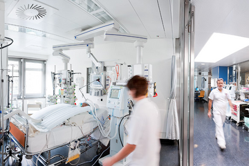 Une salle de soins intensifs. Une infirmière près d'un patient alité. Un infirmier marche dans le couloir voisin.