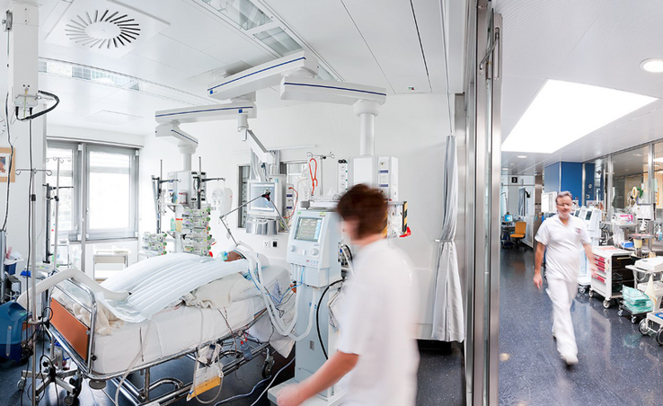 Une salle de soins intensifs. Une infirmière près d'un patient alité. Un infirmier marche dans le couloir voisin.