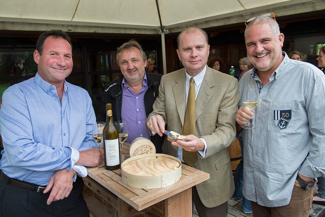 Les personnages posent autour d'une table sur laquelle est posée un gros vacherin est une bouteille de vin blanc. Monsieur Leuba tartine du vacherin sur une tranche de pain. Monsieur Gex tient un verre de vin.