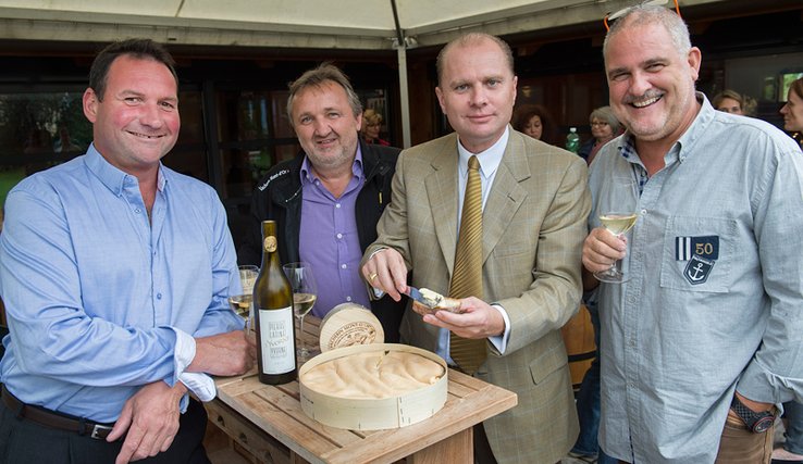 Les personnages posent autour d'une table sur laquelle est posée un gros vacherin est une bouteille de vin blanc. Monsieur Leuba tartine du vacherin sur une tranche de pain. Monsieur Gex tient un verre de vin.