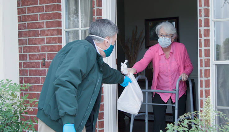 Un retraité donne un sachet repas à une personne âgée, qui est à sa fenêtre, avec un déambulateur.