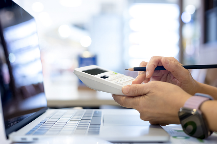 Deux mains font une addition sur une calculette, devant un écran de laptop (image d'illustration)