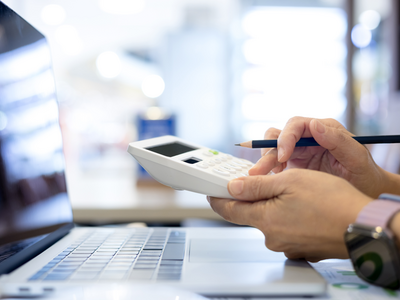 Deux mains font une addition sur une calculette, devant un écran de laptop (image d'illustration)