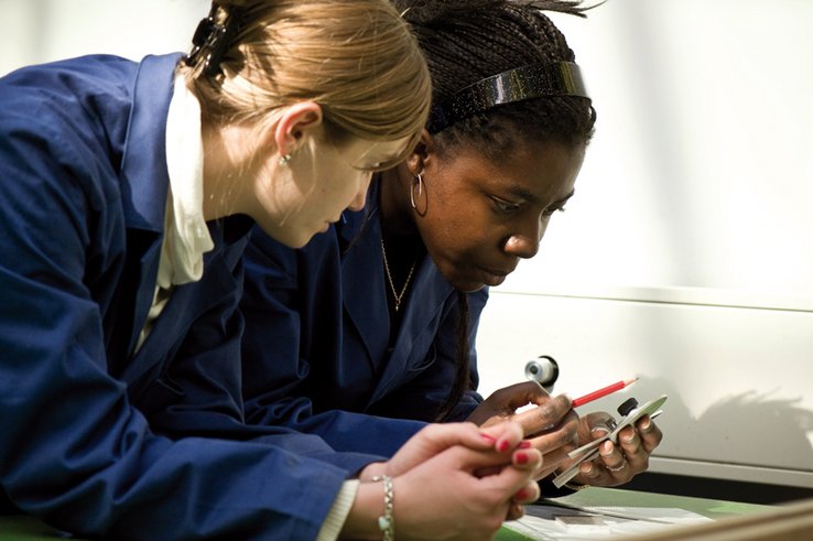 Deux apprenties techniciennes en blouse bleue, celle au second plan tient dans sa main un instrument de mesure.