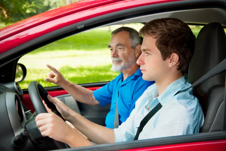 Un jeune élève conducteur au volant d'une voiture; sur le siège passager, un inspecteur ou un moniteur d'auto-école, plus âgé.