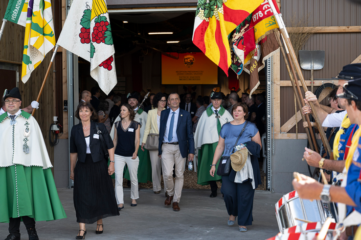 Patrick Simonin à la sortie d'une halle entouré de diverses personnalités politiques, sous une rangée de drapeaux.