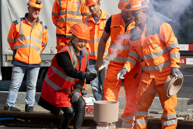 Coiffée d'un casque de chantier et vêtue d'une tenue orange comme les collaborateurs de l'entreprise de travaux publics qui l'entourent, la conseillère d'Etat tient une mèche enflammée qui va embraser l'appareil destiné à souder les rails.