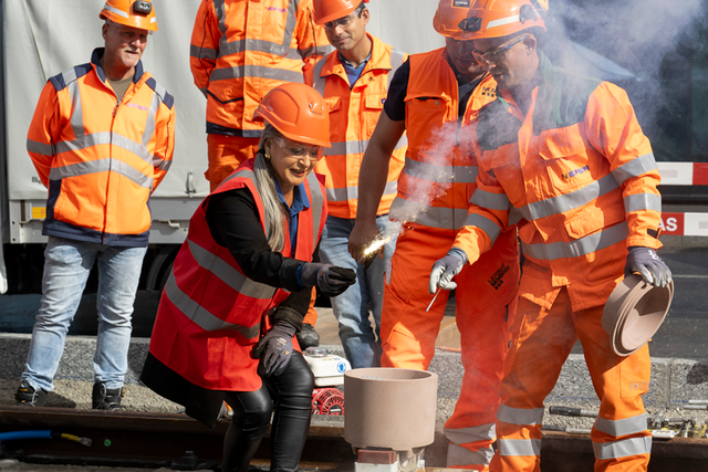 Coiffée d'un casque de chantier et vêtue d'une tenue orange comme les collaborateurs de l'entreprise de travaux publics qui l'entourent, la conseillère d'Etat tient une mèche enflammée qui va embraser l'appareil destiné à souder les rails.