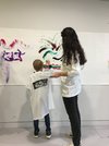 Un enfant de dos avec une blouse blanche peint; à ses côtés, une femme, de dos également, regarde.
